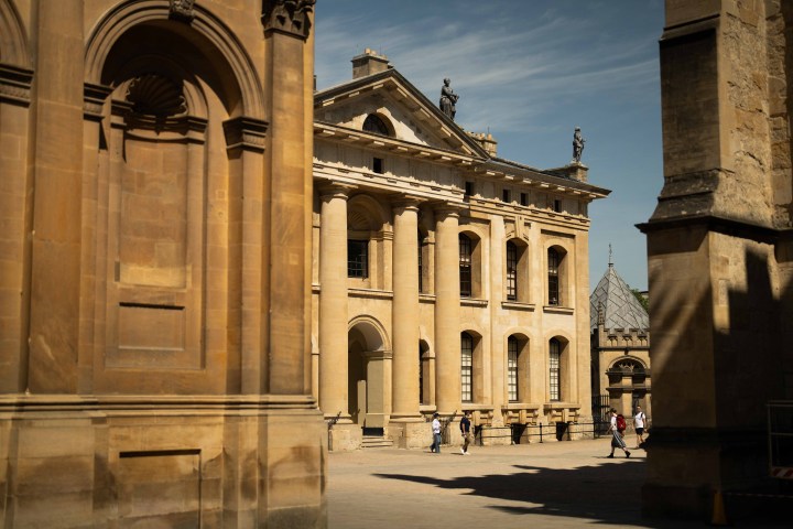 People walking near historic stone building with columns and statues under a clear sky.