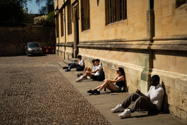 People sit against a sunlit stone wall on a cobblestone street, some reading, in a tranquil setting.