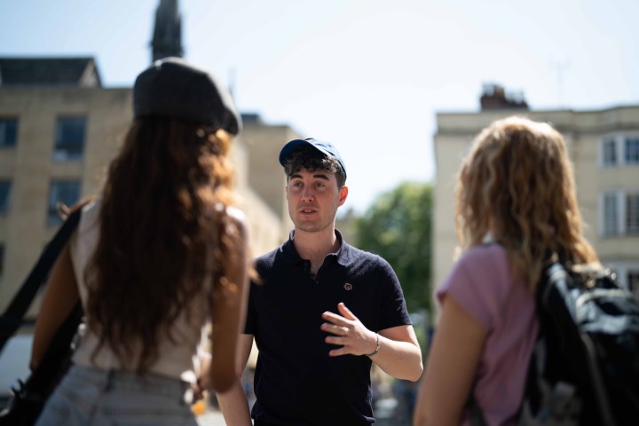 A man in a cap gestures while talking to two women on a sunny street.