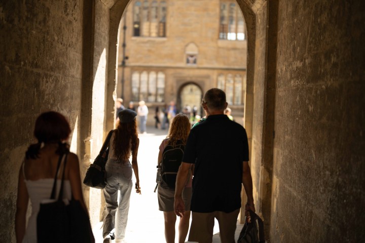 People walking through a sunlit stone archway toward a courtyard.