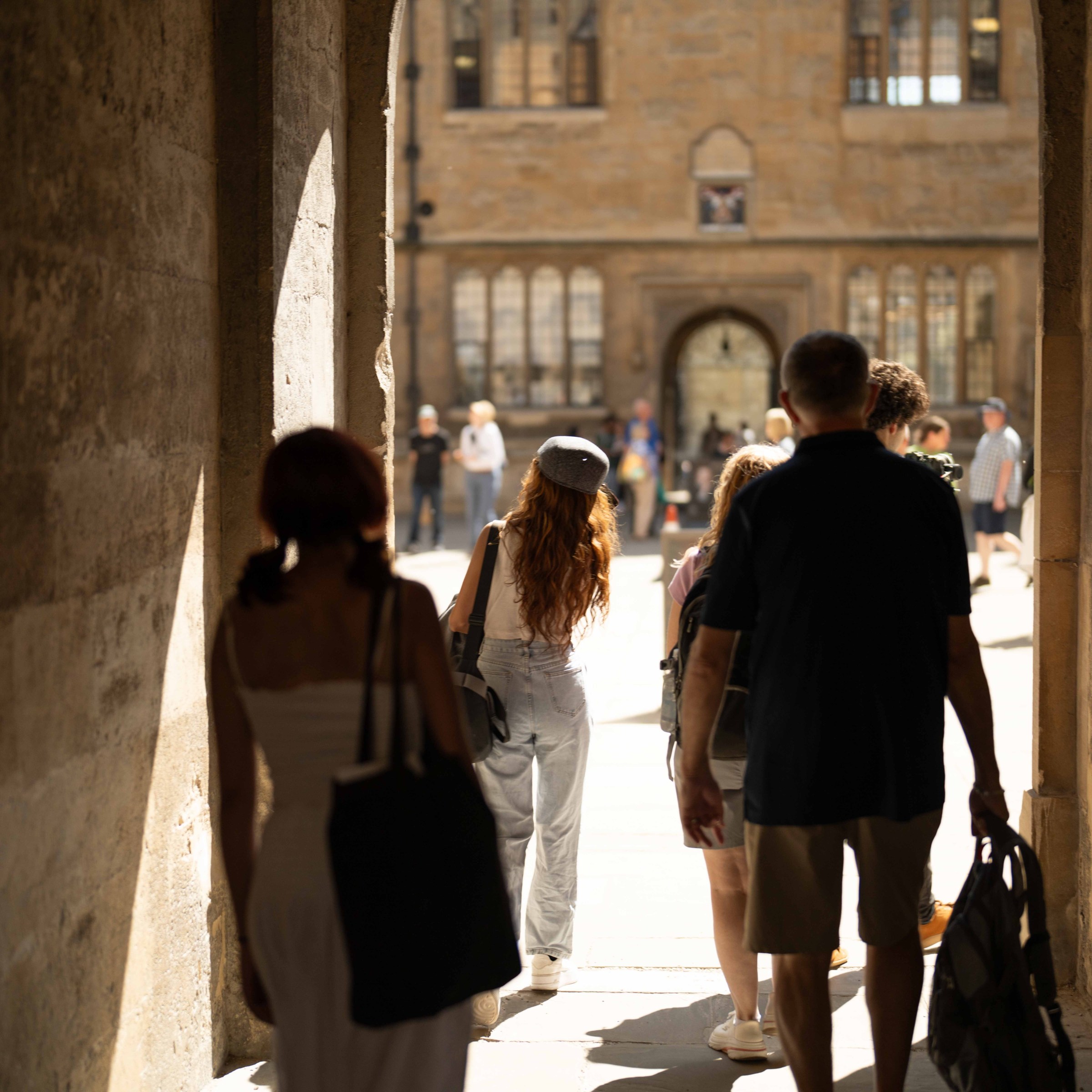 People walking through a stone archway toward a sunlit courtyard.