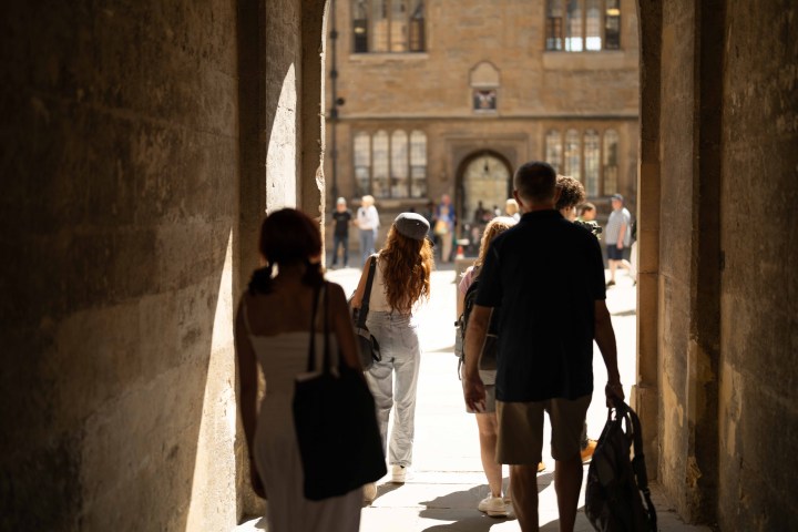 People walking through a stone archway toward a sunlit courtyard.