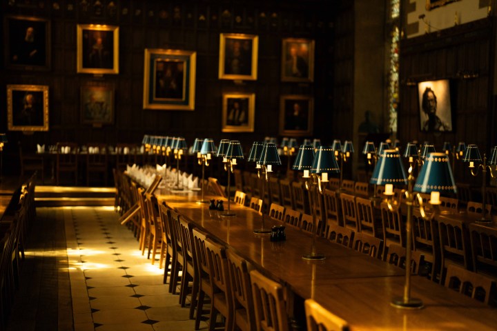 Long dining hall with wooden tables, chairs, blue lamps, and portraits on the walls.