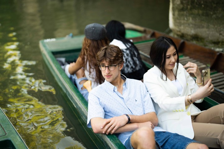 Four people in a boat, two facing the camera, one taking a selfie, on a calm water body.