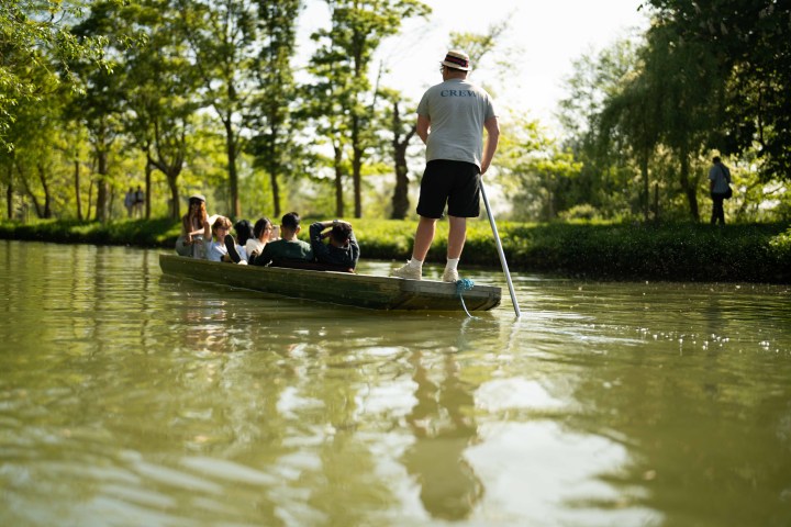 People enjoying a punt ride on a sunlit river with leafy green trees.