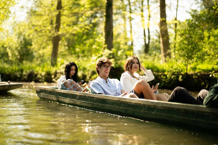 Three people relaxing in a flatboat on a sunny day surrounded by trees.