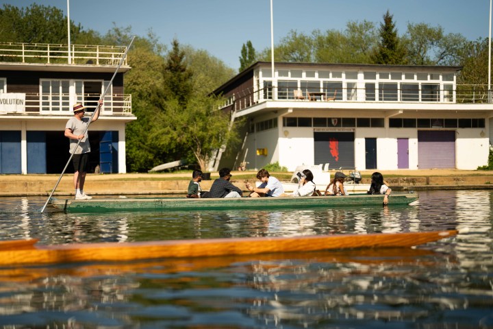 People punting on a river near two boathouses on a sunny day.