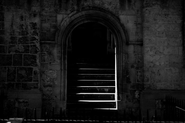 Dark stone archway with sunlit steps leading upwards in shadowy setting.